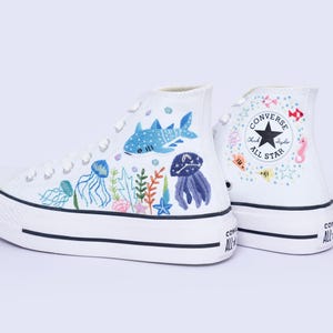May include: A pair of white high-top platform trainers featuring hand-painted marine life illustrations including a whale shark, jellyfish, seaweed, and coral. The Converse All Star logo is visible on the side with small colourful fish accents.