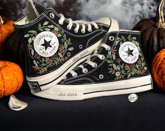 Könnte beinhalten: Ein Paar schwarze Converse-Sneaker mit hohem Schaft, die mit Blumenmustern bestickt sind und den Schriftzug "Converse All Star" an der Seite tragen. Die Schuhe stehen auf einer dunklen Oberfläche mit Kürbissen im Hintergrund.