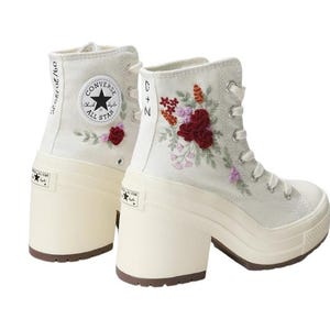 May include: Off-white high-top platform sneakers with floral embroidery. The sneakers feature a cream-colored platform sole and a lace-up closure. The Converse logo is visible on the side and heel. The embroidery includes red, pink, and purple flowers.