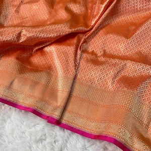 May include: A vibrant orange fabric featuring a repeating geometric diamond pattern with a metallic sheen. The textile includes a wide, intricate border with gold-toned woven motifs and a solid, bright pink trim along the bottom edge.