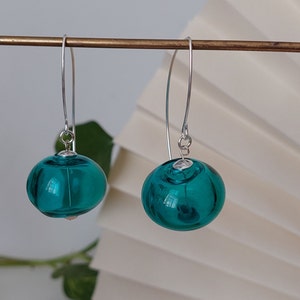 May include: A pair of teal glass ball earrings with silver-colored ear wires. The earrings are round and have a reflective surface. The earrings are hanging from a gold-colored bar.