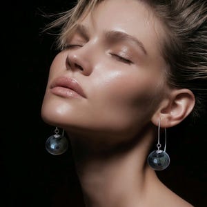 May include: A pair of silver drop earrings featuring smooth, spherical dark gray glass beads suspended from thin, curved silver wires. The earrings hang vertically, showcasing a polished metallic finish and a reflective, translucent orb design.