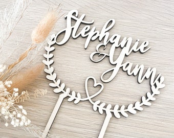 May include: A wooden cake topper with the names "Stephanie & Yann" in elegant script. The names are within a circular wreath design with a heart detail. The topper has two pointed stakes for insertion into a cake.
