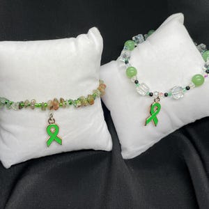 May include: Two green ribbon charm bracelets with green and brown stone beads. The bracelets are on white pillow-like surfaces.
