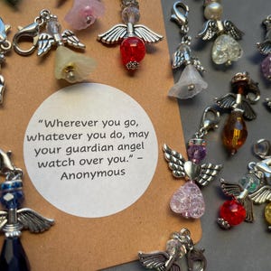 Puede incluir: Una colección de dijes de ángel de plata con cuentas de vidrio de colores y cristales. Los dijes están unidos a llaveros y tienen una cita sobre ángeles guardianes.