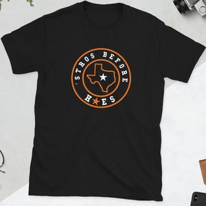 Könnte beinhalten: Schwarzes T-Shirt mit einem kreisförmigen orange-weißen Grafikdesign. Das Design zeigt den Umriss von Texas mit einem Stern in der Mitte und den Worten "STROS BEFORE H*ES" am Rand. Das T-Shirt hat kurze Ärmel.