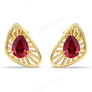Puede incluir: Un par de pendientes de oro. Cada pendiente presenta una piedra preciosa roja oscura en forma de lágrima engastada en un diseño de rayos de sol estilizado. Los pendientes se presentan sobre un fondo blanco.