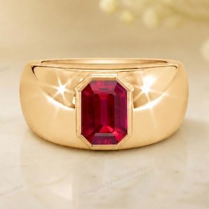 Può includere: Un anello d'oro con una grande pietra preziosa rosso scuro rettangolare al centro. L'anello ha una fascia liscia e arrotondata e una finitura lucida, che riflette la luce. La pietra preziosa è incastonata in una lunetta rettangolare.