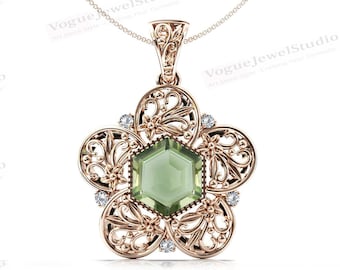 Può includere: Una collana con un ciondolo in oro rosa decorato con una pietra preziosa verde di forma esagonale, circondata da un delicato motivo a filigrana. Il ciondolo è attaccato a una catena in oro rosa.
