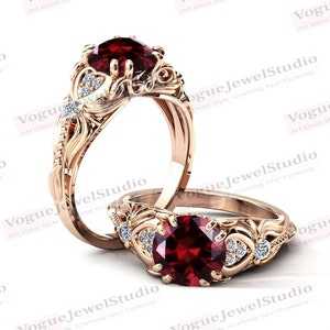 Op de afbeelding: Twee roségouden ringen met een grote, ronde, dieprode edelsteen. De ringen hebben ingewikkelde bloemen- en scrollwerkontwerpen, geaccentueerd met kleine, heldere edelstenen. De ringen zijn van Vogue JewelStudio.