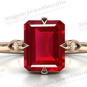 Puede incluir: Un anillo de oro rosa con una gran piedra preciosa de rubí de talla esmeralda y dos pequeños acentos de diamantes a cada lado. El anillo tiene un diseño delicado de inspiración Art Deco.