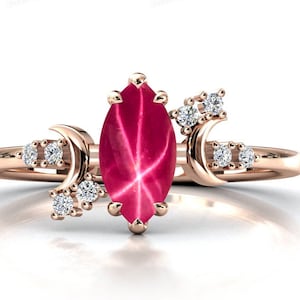 May include: A rose gold ring with a large, red star ruby gemstone and two crescent moon accents set with smaller diamonds.