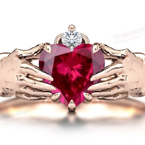 Może przedstawiać: Złoty pierścionek z dwoma rękami trzymającymi duży, sercowaty rubin. Mniejszy diament znajduje się na górze rąk.