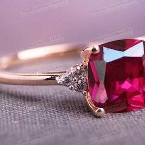 May include: A rose gold ring with a large, square-shaped, deep red gemstone surrounded by smaller, clear gemstones.
