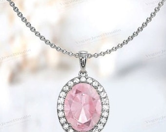 May include: A silver necklace with an oval pink gemstone pendant. The pendant is surrounded by a halo of small, clear gemstones. The chain is delicate and silver, and the background is blurred.