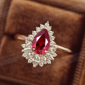 Pode incluir: Um anel de ouro com uma gema de rubi em forma de pêra rodeada por um halo de diamantes menores.