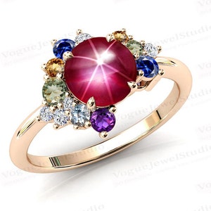 Op de afbeelding: Een ring van roségoud met een grote rode ster-robijn als steen, omringd door kleinere edelstenen in verschillende kleuren, waaronder groen, blauw, paars en wit.