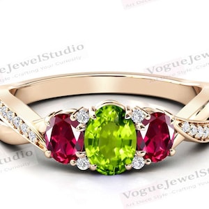 Könnte beinhalten: Ein goldener Ring mit einem gedrehten Band und drei ovalen Edelsteinen. Der mittlere Stein ist ein leuchtend grüner Peridot, und die beiden seitlichen Steine sind tiefrote Rubine. Das Band ist mit kleinen, klaren Diamanten verziert.
