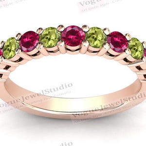 May include: A rose gold band ring with alternating green and red gemstones. The ring is engraved with the text "Vogue Jewel Studio Art Deco Style Crafting Your Curiosity".
