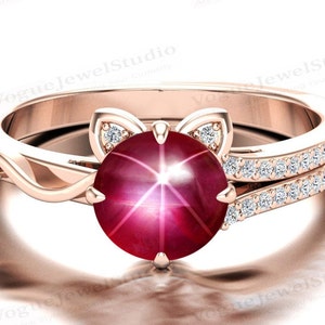 May include: A rose gold ring featuring a large red star ruby gemstone surrounded by smaller diamonds. The ring has a delicate cat-shaped design with diamond accents on the ears.
