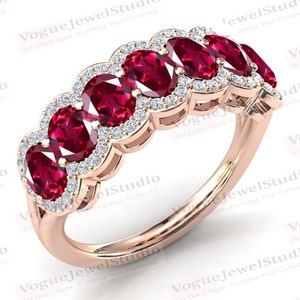 Puede incluir: Un anillo de oro rosa con una serie de piedras preciosas rojas oscuras de corte ovalado, cada una rodeada por un halo de pequeños diamantes brillantes. El anillo lleva la inscripción "Vogue JewelStudio".