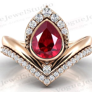 May include: A pear-shaped ruby gemstone ring set in a rose gold band with a diamond halo and a matching diamond band. The ring features a V-shaped design with a pointed tip.