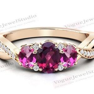 May include: A rose gold ring with a twisted band and three oval-shaped gemstones. The center stone is a large, deep pink gemstone, flanked by two smaller pink gemstones. The band is set with small, clear diamonds.