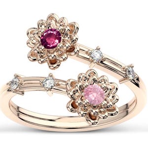 May include: A rose gold ring with two flower-shaped settings, each with a pink gemstone. The ring is adorned with smaller clear gemstones.