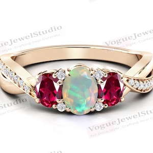 以下が含まれることがあります： ねじれたバンドと3つの楕円形の宝石が付いた金の指輪。中央の石は白いオパールで、両側の石は赤いルビーです。指輪には小さなダイヤモンドが埋め込まれています。