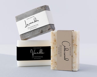 May include: Three bars of handmade soap with labels. The soap on the left is white with a black label that says "Vanilla 100% Handmade Soap". The soap in the middle is gray with a white label that says "Lavender 100% Handmade Soap". The soap on the right is white with brown specks and a brown label that says "Cedarwood 100% Handmade Soap".