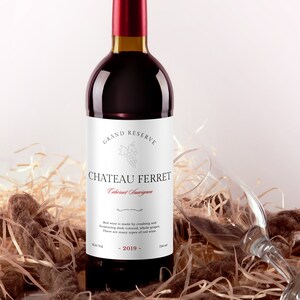 May include: A bottle of red wine with a white label that reads "GRAND RESERVE CHATEAU FERRET Cabernet Sauvignon". The label also includes a small image of grapes and the text "Red wine is made by crushing and fermenting dark-colored, whole grapes. There are many types of red wine." The bottle is sitting on a bed of straw.