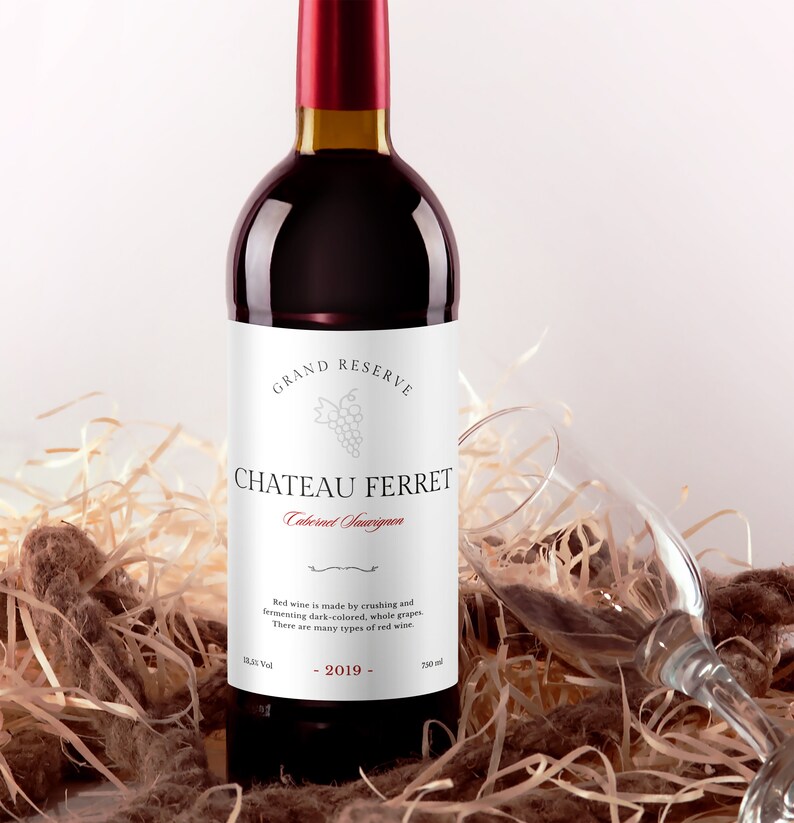 May include: A bottle of red wine with a white label that reads "GRAND RESERVE CHATEAU FERRET Cabernet Sauvignon". The label also includes a small image of grapes and the text "Red wine is made by crushing and fermenting dark-colored, whole grapes. There are many types of red wine." The bottle is sitting on a bed of straw.