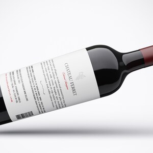 May include: A bottle of Chateau Ferret Cabernet Sauvignon wine with a white label and a red cap. The label features the winery's name and a cluster of grapes. The bottle is lying on a white surface.