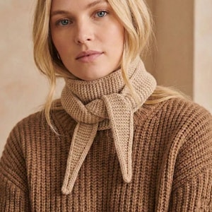 May include: A beige knitted scarf tied around the neck, with the ends hanging down. The scarf is made of a thick, textured yarn. The person is wearing a brown, ribbed knit sweater. The overall look is warm and cozy.