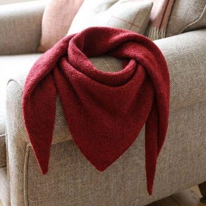 Può includere: Uno scialle triangolare lavorato a maglia, di colore rosso intenso, drappeggiato su una poltrona dai toni neutri. La trama dello scialle è visibile, con estremità a punta. Lo sfondo presenta cuscini in morbide tonalità rosa e beige, suggerendo un'atmosfera accogliente.