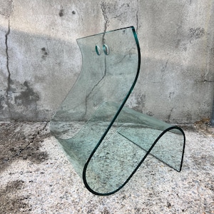 May include: A clear glass chair with a curved, S-shaped design. The chair has two small holes on the back.