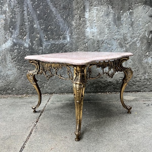 May include: A square table featuring a light pink marble top with beveled edges. The base is constructed from ornate, gold-toned metal with intricate scrollwork and four curved legs that terminate in small, rounded feet at the bottom.