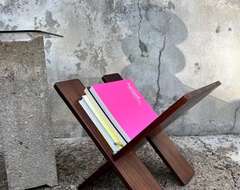 May include: A wooden magazine rack with a criss-cross design, holding a stack of books, including a bright pink book with the title "DESIGN" on the cover.