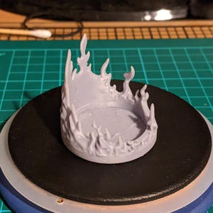 Puede incluir: Una base de miniatura impresa en 3D de color púrpura claro con forma de anillo de llamas. La base está diseñada para ser utilizada en juegos de mesa o para pintar miniaturas.