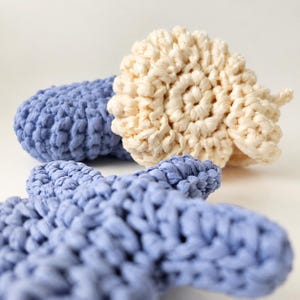 May include: A set of crocheted items featuring a cream-colored spiral shell shape and two periwinkle blue starfish shapes. The objects are crafted from thick yarn with a visible knotted texture, showcasing a distinct handmade construction style.