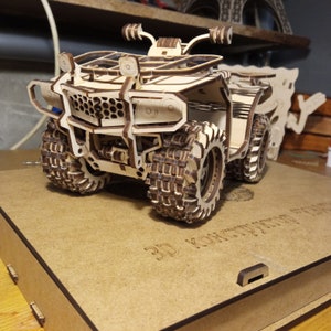 以下が含まれることがあります： 詳細なデザインのATV（全地形対応車）の木製モデル。モデルは組み立て済みで、展示の準備ができています。モデルは、「3D Конструктор」という文字が印刷された木製台座の上に置かれています。