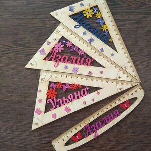 May include: Three wooden protractors with pink, purple, and yellow butterflies and flowers. The protractors are labeled with the names "Azalia", "Uljana", and "Azalia".