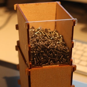 Può includere: Una pila di tre scatole di legno con un coperchio di plastica trasparente. La scatola superiore è piena di piccole viti argentate.