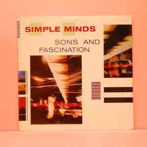 Peut inclure: Pochette de l'album "Sons and Fascination" de Simple Minds. La couverture est blanche avec le nom du groupe en rouge et le titre de l'album en noir. La couverture présente des images abstraites et un fond rose.