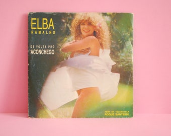 Puede incluir: Una portada de disco de vinilo vintage con una mujer de cabello largo y rubio que lleva un vestido blanco. La portada está titulada "Elba Ramalho" y presenta el título de la canción "De Volta Pro Aconchego". El texto "Tema da Telenovela Roque Santeiro" también está incluido en la portada.