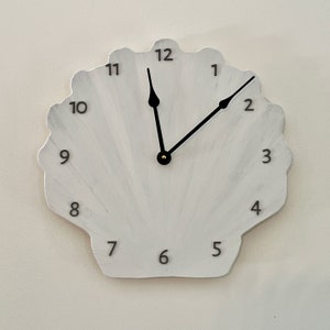 May include: White seashell-shaped wall clock with black numerals and hands. The clock has a light gray and white painted finish. The clock hands point to approximately 1:08. The clock is mounted on a light-colored wall.