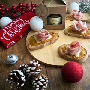 Op de afbeelding: Een feestelijke kerstarrangement met geroosterde broodjes belegd met kaas en prosciutto, een pot marmelade en decoraties. Inclusief dennenappels, kerstballen en een rode handdoek met de tekst "Merry Christmas". De opstelling staat op een houten plank.