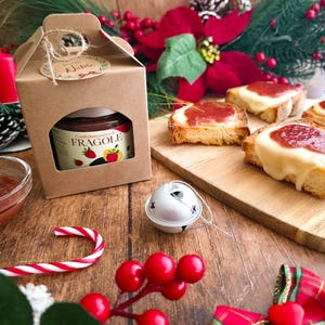 Op de afbeelding: Een pot aardbeienjam in een kartonnen geschenkdoos, met een venster dat het etiket laat zien. De afbeelding bevat toast met jam en kaas, een zuurstok en kerstversieringen. Het etiket luidt "Fragole".