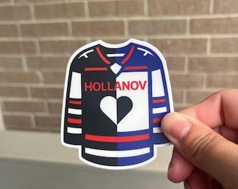 Puede incluir: Una pegatina con forma de camiseta de hockey. La camiseta está dividida verticalmente, una mitad negra y la otra blanca. Una forma de corazón está en el centro. La camiseta tiene detalles rojos y azules y la palabra "HOLLANOV".