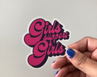 Op de afbeelding: Een roze en zwarte sticker met de tekst "Girls support Girls" in een retro-lettertype.